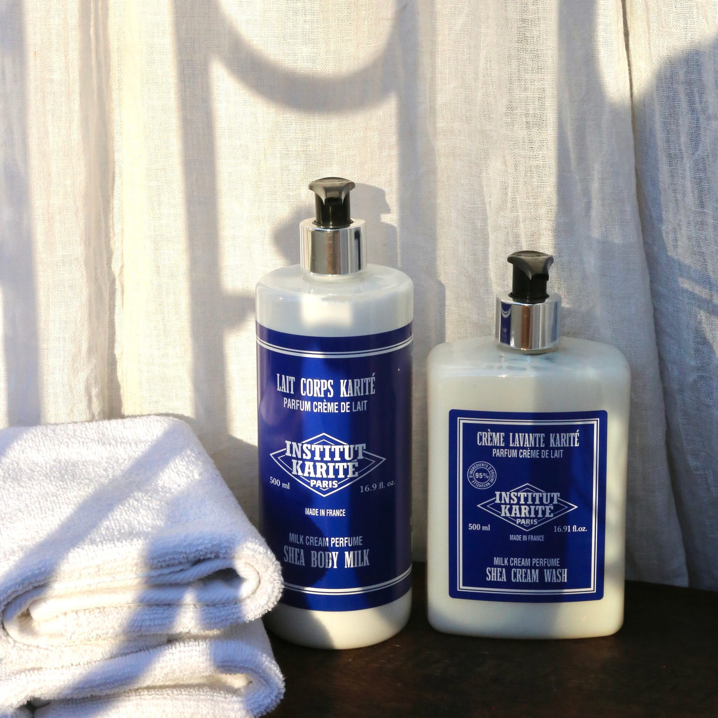 Shea Cream Wash 500ml bottle beside Shea Body Milk 500ml with folded towels against linen