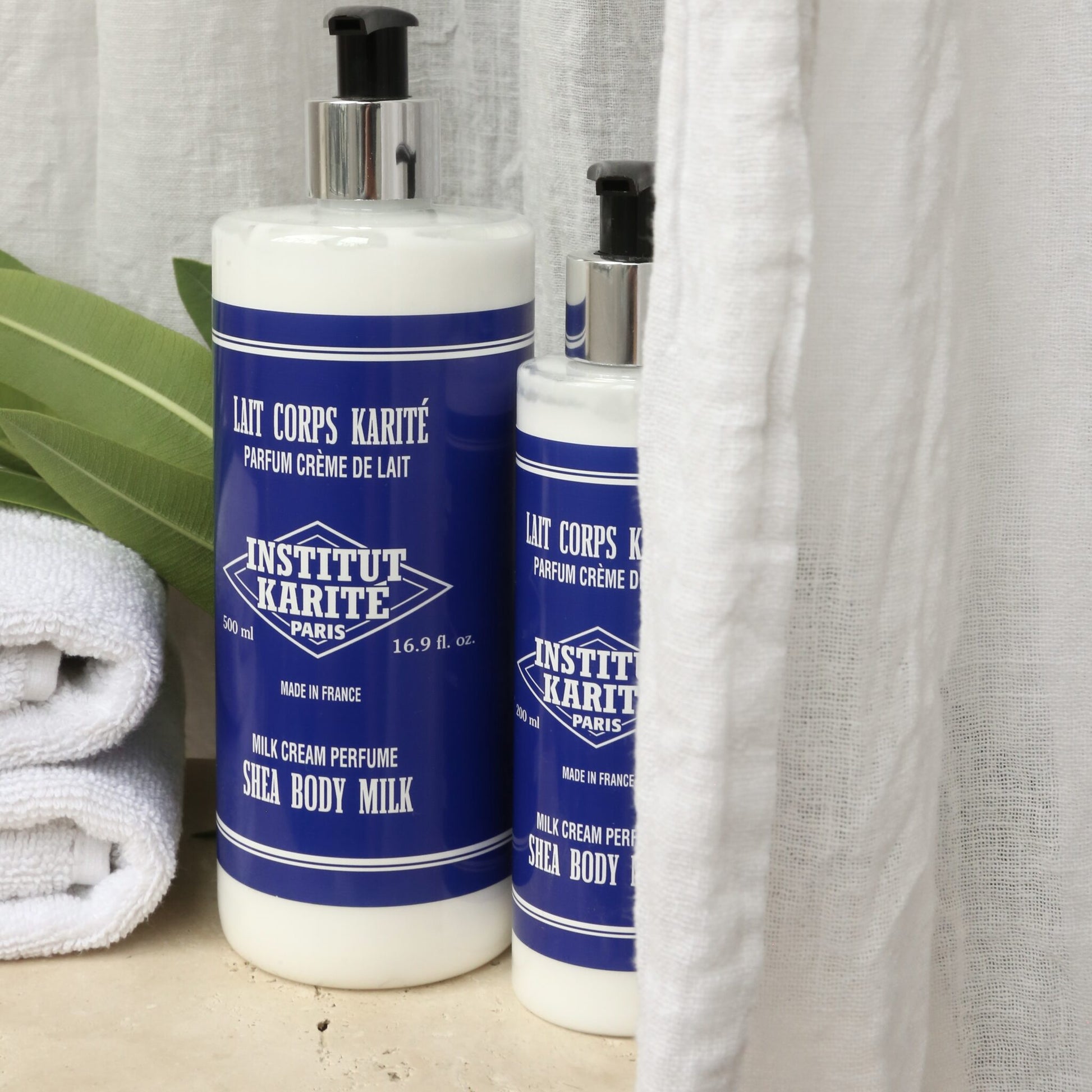 500mL and 200mL Milk Cream Shea Body Milk by Institut Karité Paris against a linen curtain with folded towels