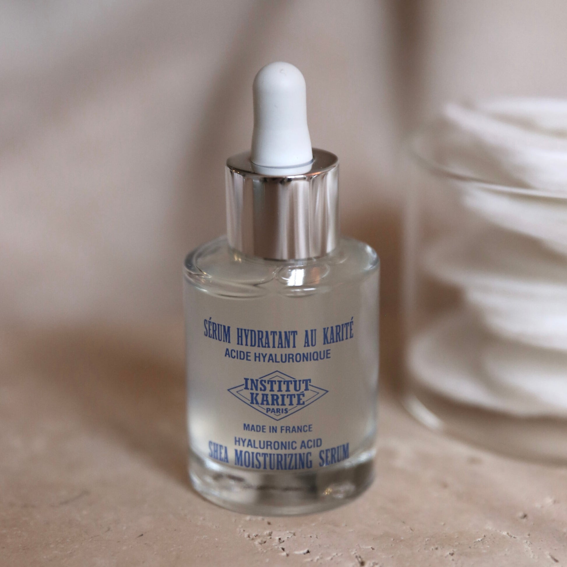 Close view of the Shea Moisturising Serum bottle on stone beside a jar of cotton pads