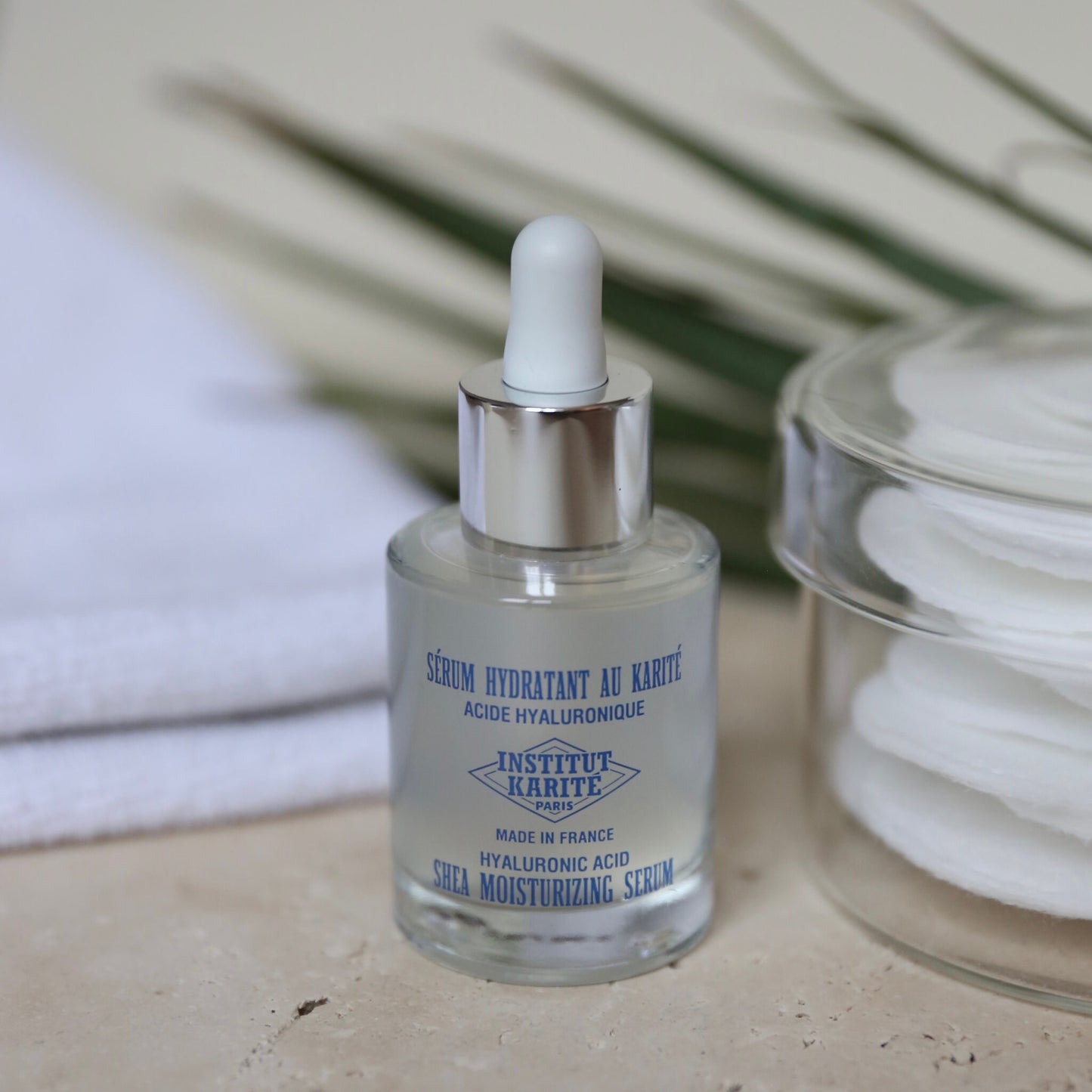 Shea Moisturising Serum bottle on stone with cotton pads, a folded towel and palm leaves