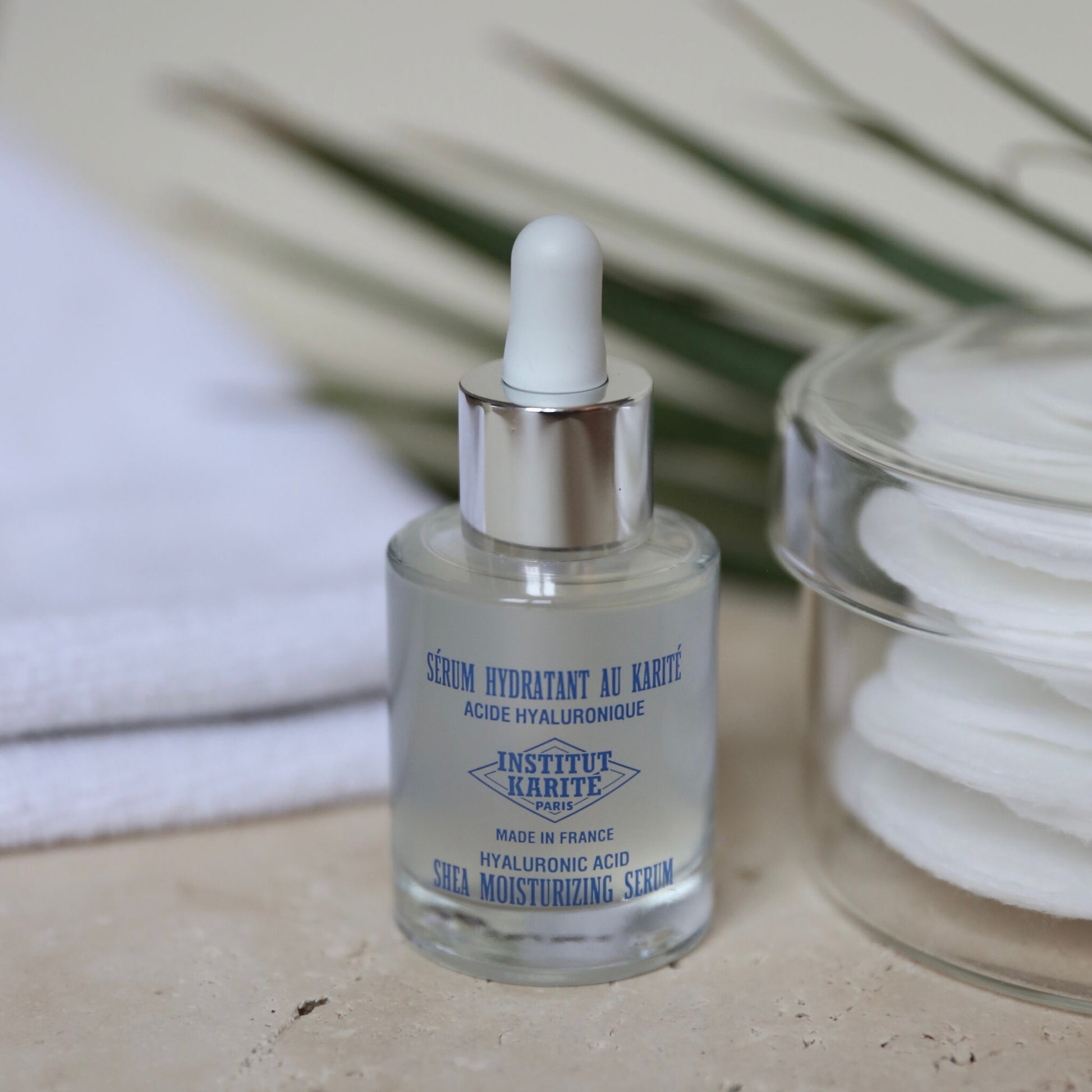 Shea Moisturising Serum bottle on stone with cotton pads, a folded towel and palm leaves