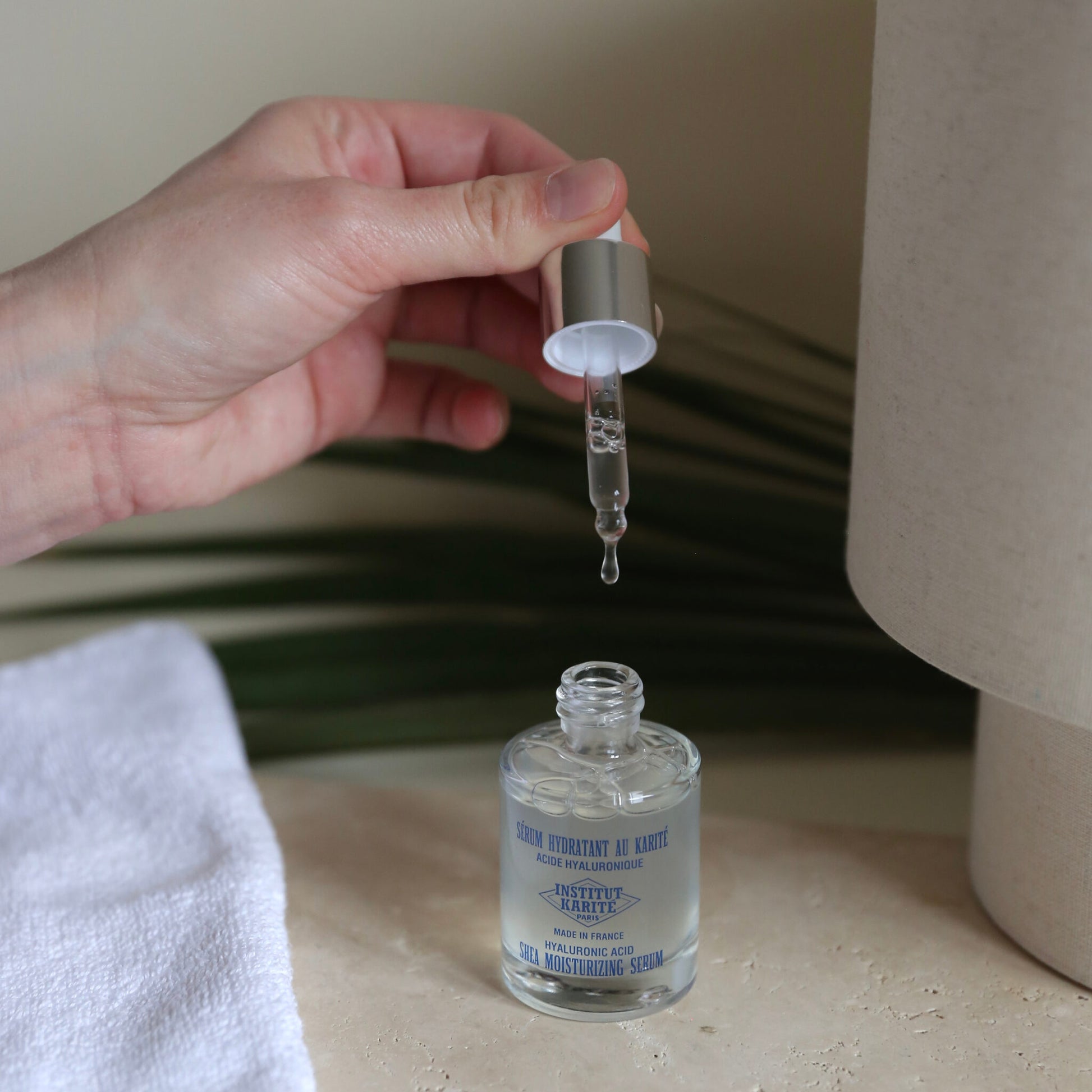 Hand holding the Shea Moisturising Serum dropper above the open bottle with a drop falling