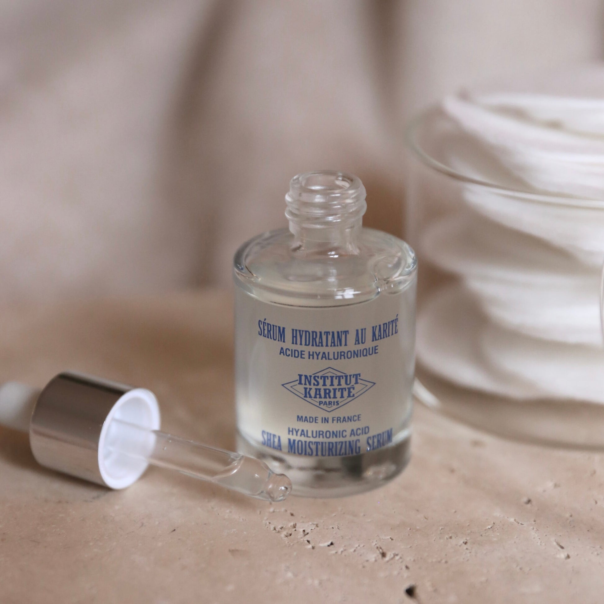 Open bottle of Shea Moisturising Serum with its dropper resting on stone beside cotton pads