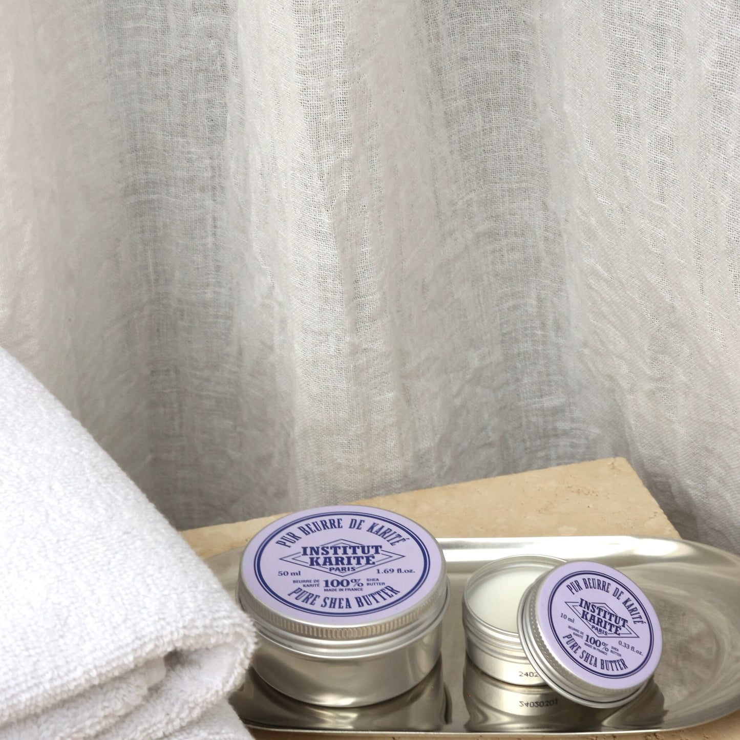 150 ml and travel size tins of Institut Karité Paris Pure Shea Butter on a metal tray beside a folded white towel