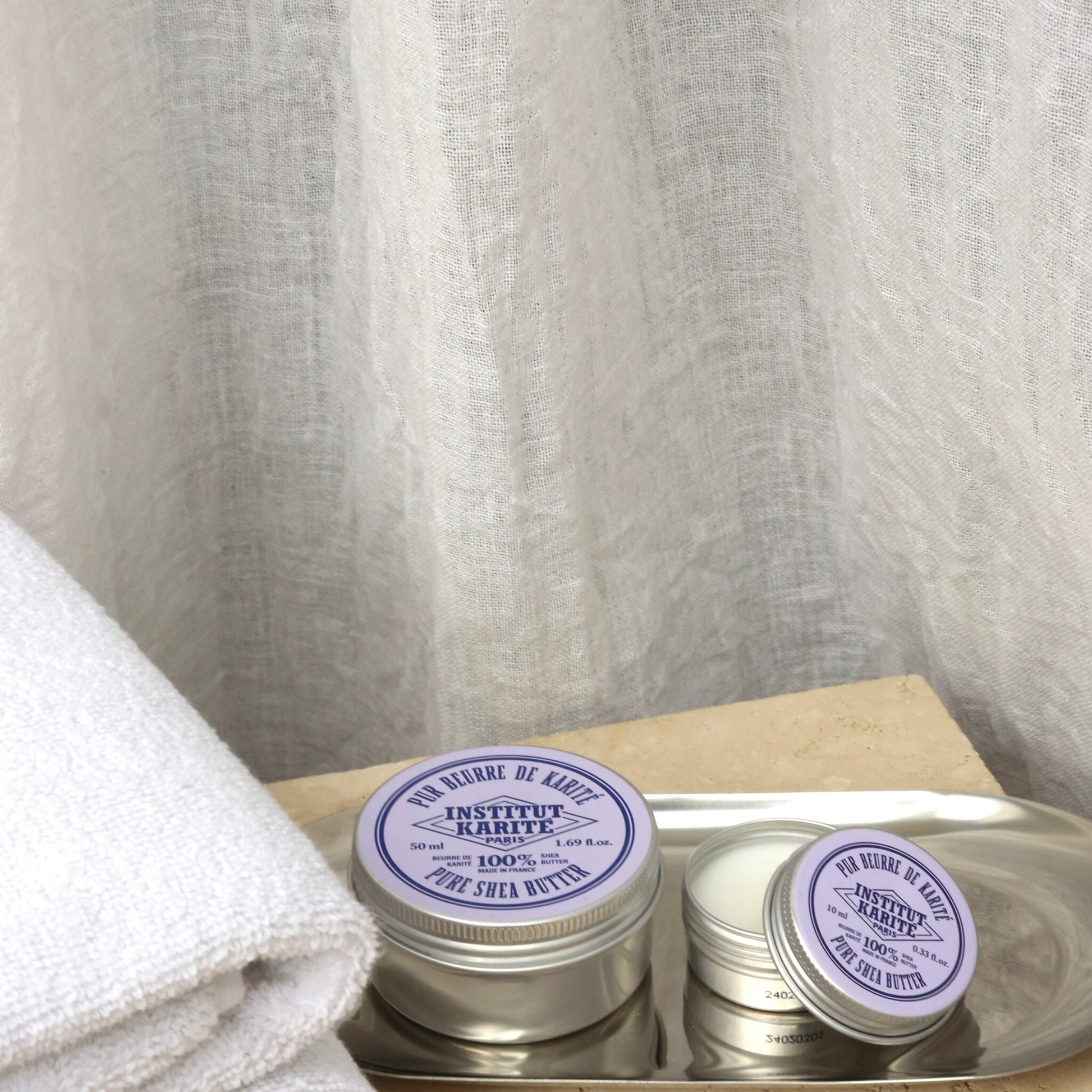 150 ml and travel size tins of Institut Karité Paris Pure Shea Butter on a metal tray beside a folded white towel
