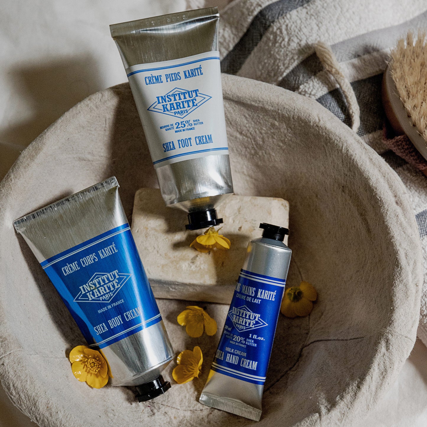 Shea Foot Cream, Shea Body Cream and Shea Hand Cream tubes in a stone bowl with buttercups and linen