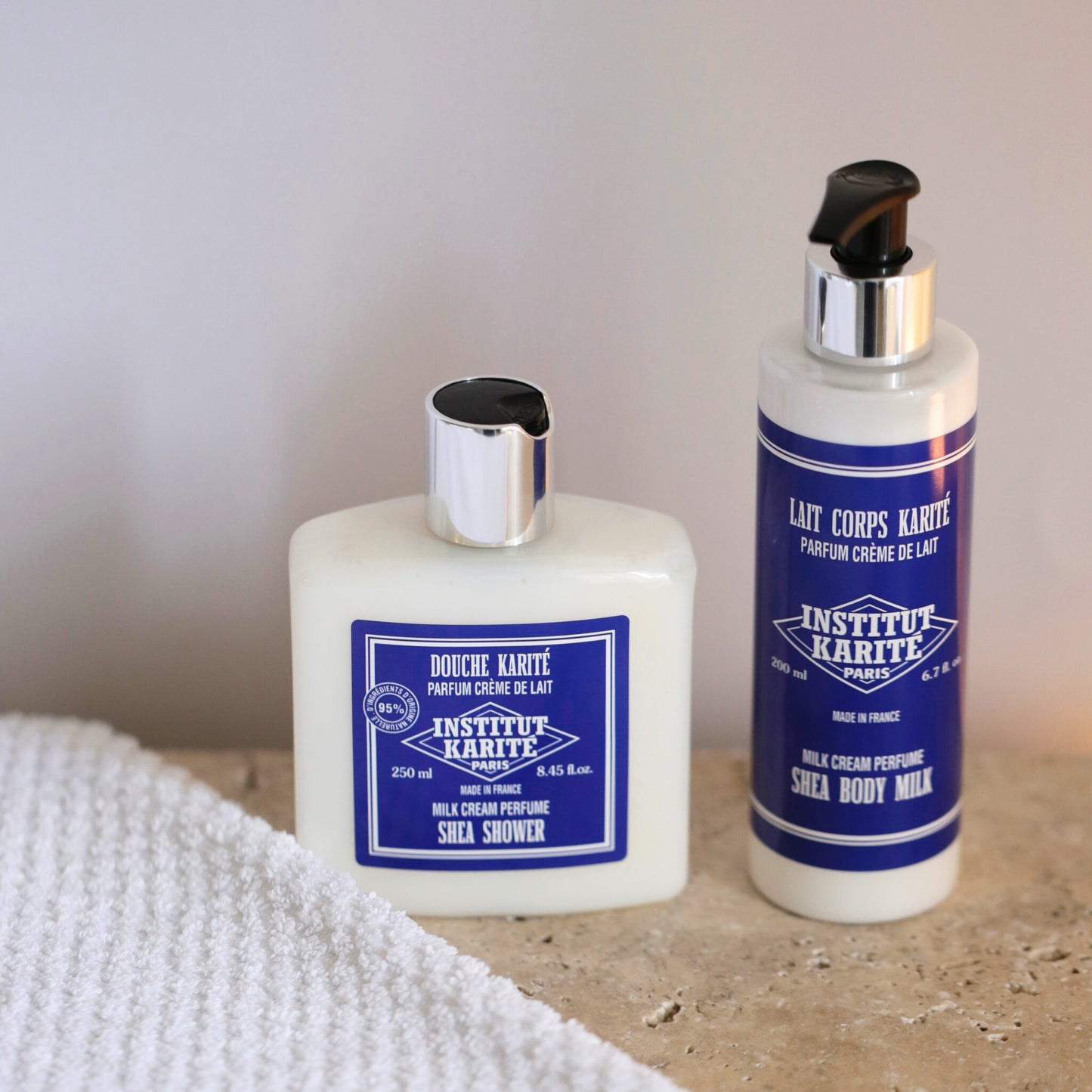 Shea Shower Milk Cream 250ml bottle beside Shea Body Milk on a stone surface with a folded towel