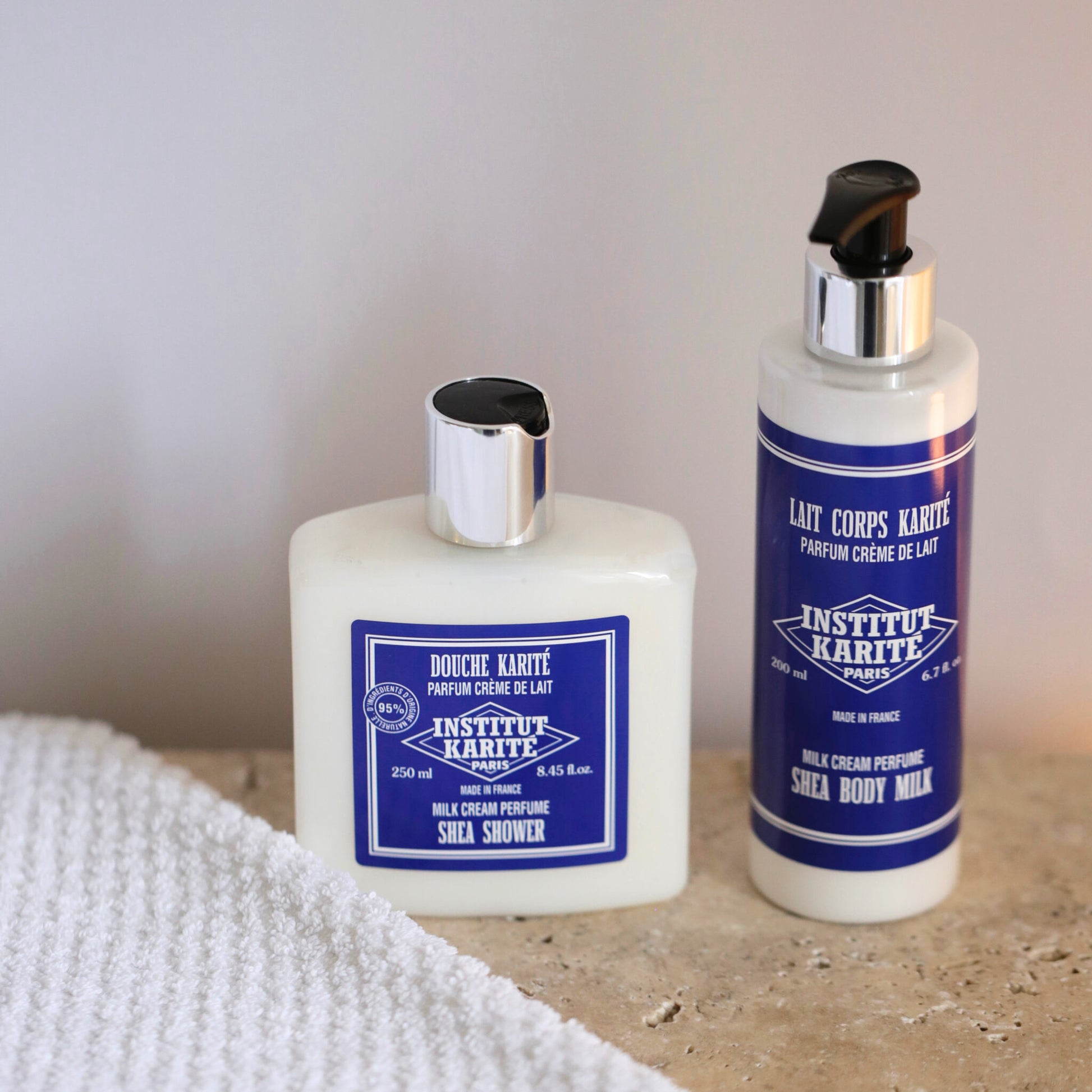 Shea Shower Milk Cream 250ml bottle beside Shea Body Milk on a stone surface with a folded towel