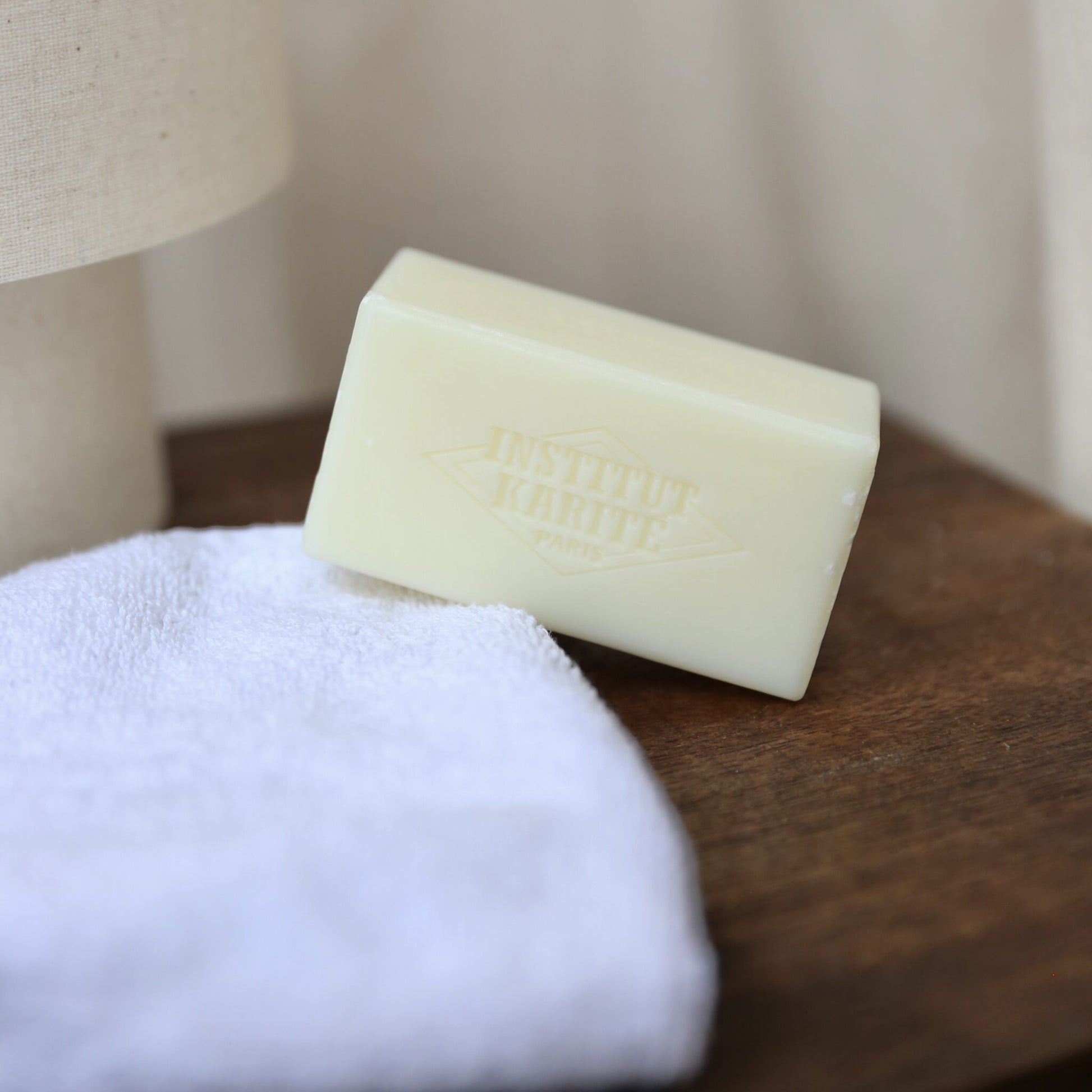 Unwrapped 150g shea soap bar by Institut Karité Paris resting on a folded white towel beside a linen lamp